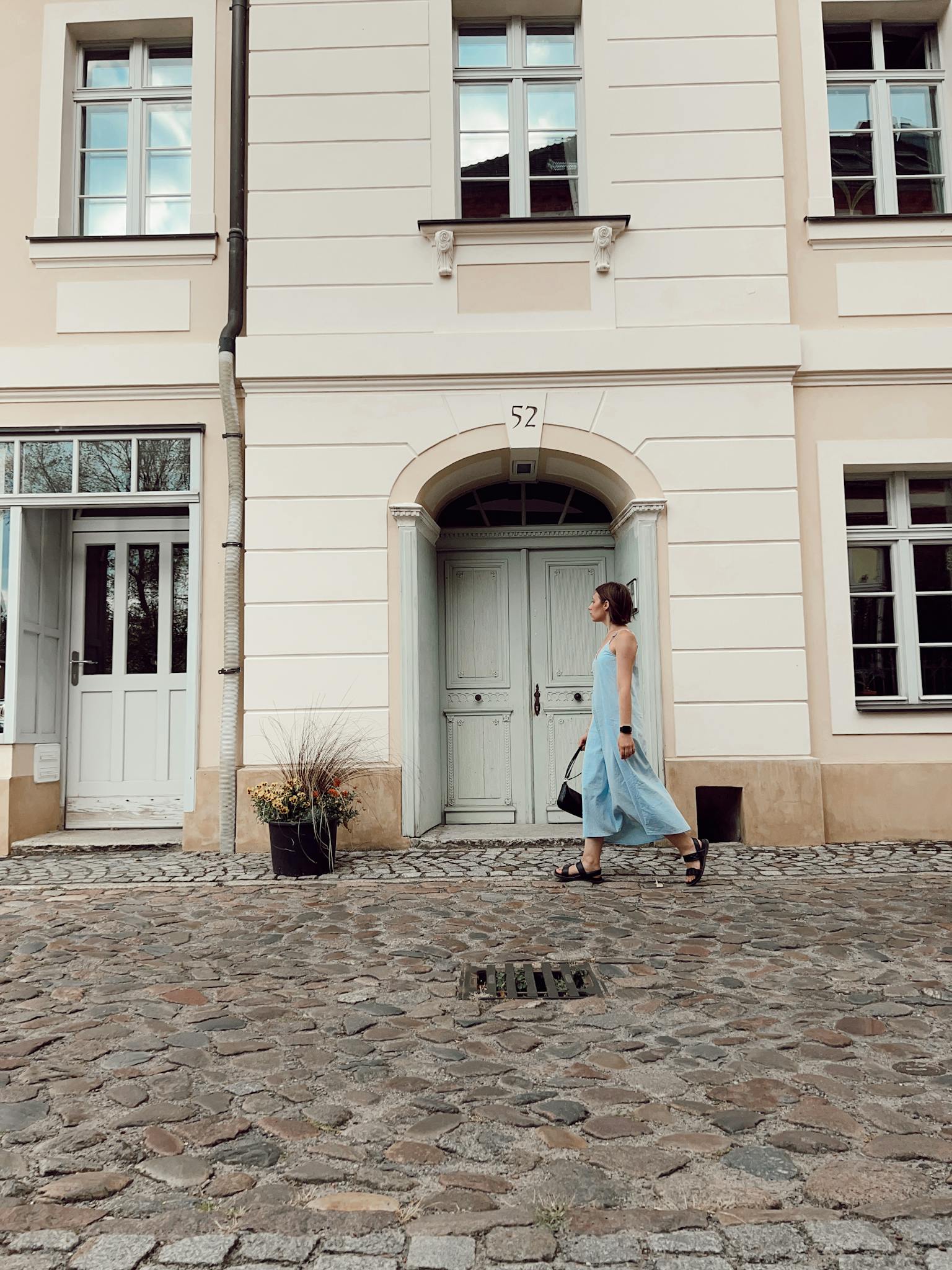 A woman in a pastel blue dress walks past a historic townhouse facade on a cobblestone street.
