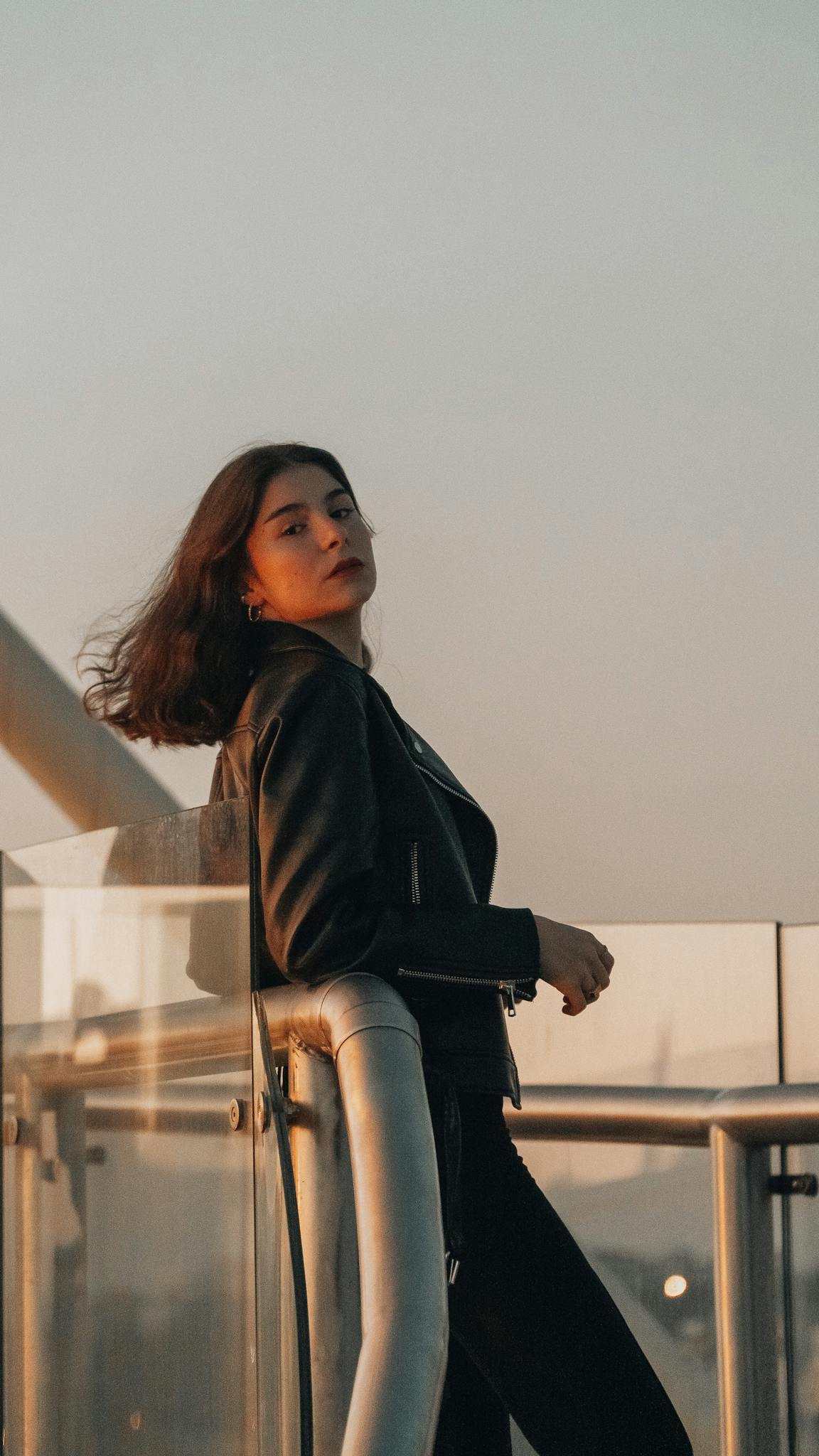 Portrait of a fashionable woman with brunette hair wearing a jacket, standing on a balcony at sunset.